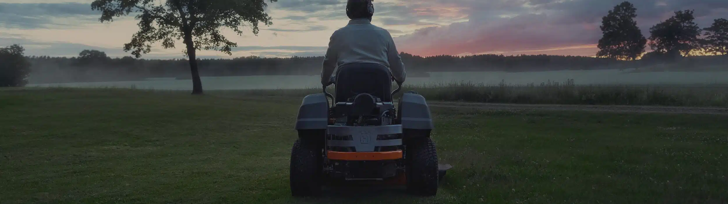 A wide banner photo of a silhouetted person driving a ride-on mower across an open field at sunset.