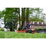 A man rides an orange zero-turn mower at speed across a large lawn with trees and a building in the background.