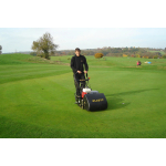 A man pushes a black Allett cylinder mower across a golf course green with autumn trees in the distance.
