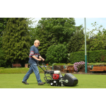 A man pushes a petrol-powered cylinder mower with a Honda engine across a bowling green, with benches and trees behind.