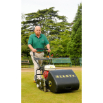 A man in a green polo shirt pushes a black Allett cylinder mower across a bowling green with trees in the background.