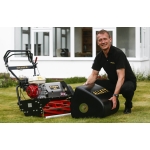 A man kneels on a lawn beside a black Allett cylinder mower in front of a house, smiling at the camera.