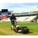 A man mows the outfield of a cricket stadium with an Allett cylinder mower.