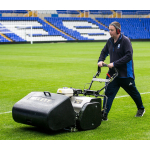 A groundsman wearing ear defenders pushes a green cylinder mower across a football stadium pitch.