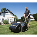 A man pushes a black Allett cylinder mower across a lawn in front of a white house under a clear blue sky.
