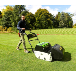 A man pushes a black and green Allett cylinder mower with a grass collection box across a striped lawn surrounded by trees.