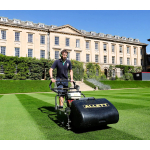 A groundskeeper pushes a black Allett cylinder mower across a striped lawn in front of a grand stone college building.