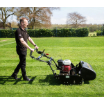A man pushes a black petrol cylinder mower across a large grass field bordered by a hedge and bare trees.