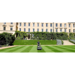A groundskeeper mows a striped lawn in front of an ornate stone building covered in trimmed ivy hedges.