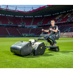 A man rides a cylinder mower fitted with a trailing seat while cutting the grass pitch inside a football stadium.