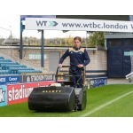 A groundskeeper in a blue tracksuit pushes a blue Allett cylinder mower across a football stadium pitch.