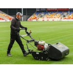 A groundsman pushes a cylinder mower across a football stadium pitch.