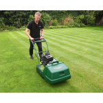 A man in a black polo shirt pushes a green cylinder lawn mower across a neatly striped garden lawn bordered by hedges.