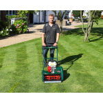 A man stands behind a dark green petrol cylinder mower on a striped lawn in front of a house with flower beds.
