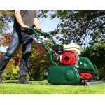 A man pushes a green cylinder mower across a lawn with autumn-coloured trees behind him under a blue sky.