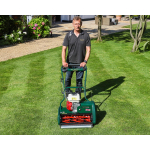 A man stands behind a green Allett cylinder mower on a neatly striped lawn, with flower beds and a house in the background.