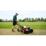A man pushes a black petrol cylinder mower along the edge of a large sports pitch with trees in the distance.