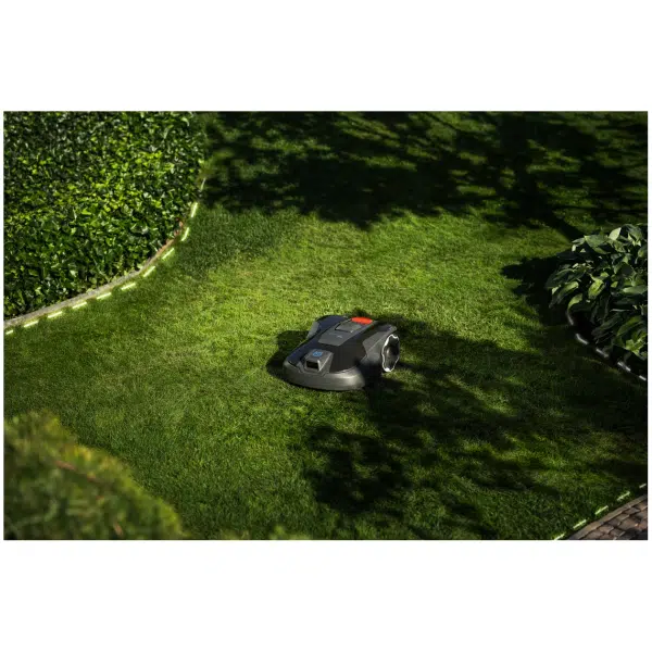 A dark robotic lawnmower with a red top panel sits on a garden lawn beside a hedge and flower bed, seen from above.
