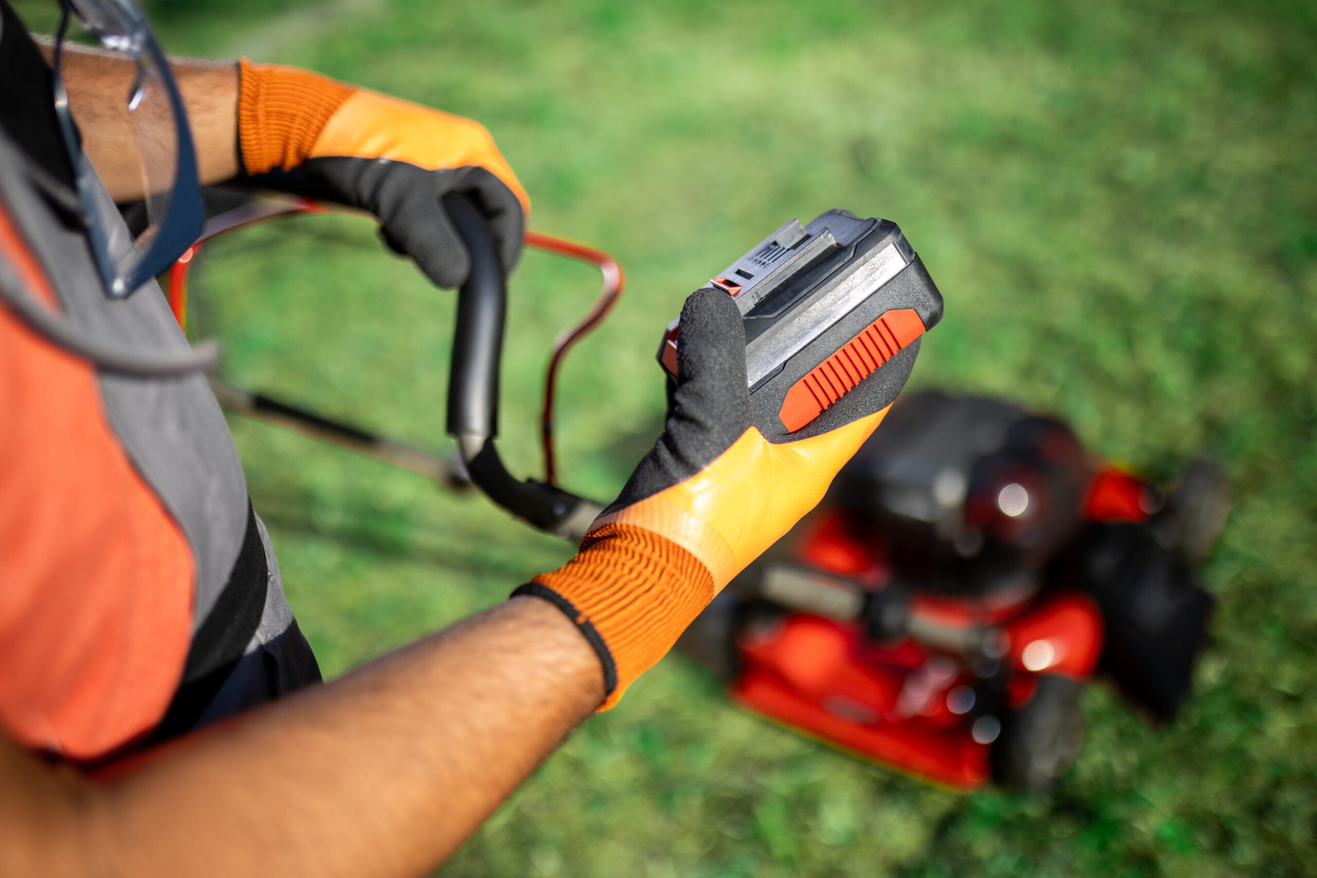 An individual holding a battery, operates a electrical lawn mower, wearing gloves and protective gear, while focusing on maintaining a well-groomed garden. Bright sunlight enhances the vibrant green grass.