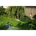 Robotic lawn mower operating in a garden with a blurred person in the background.