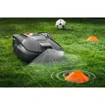 Robotic lawn mower operating in a garden with a soccer ball and cone nearby.